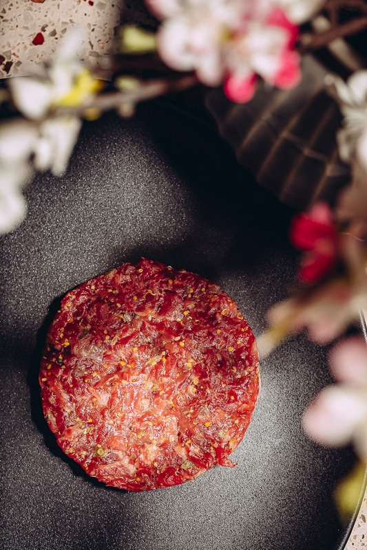 A disc of hand-cut beef tartare on dark stone, blossom in the background.