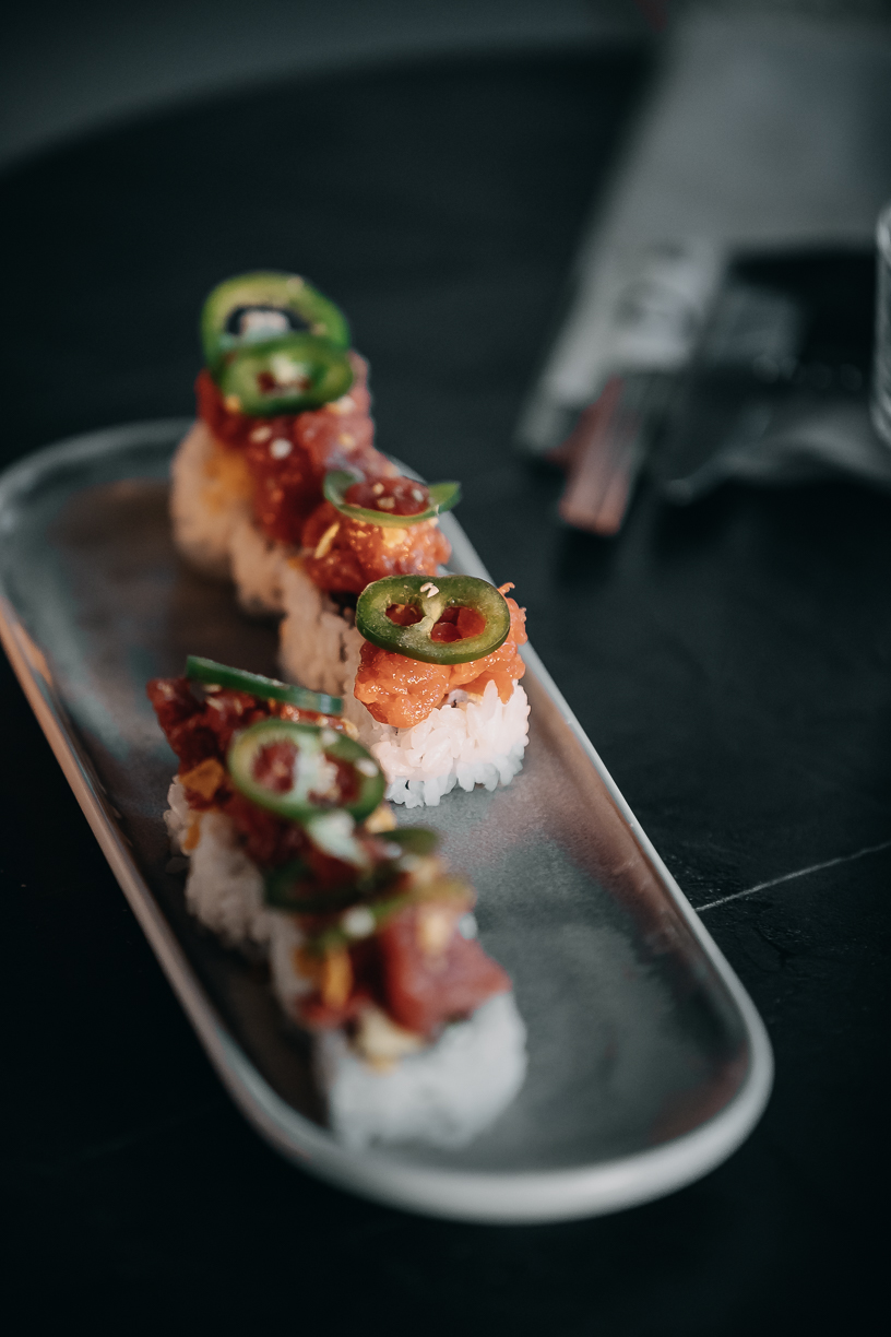A row of beef sushi topped with jalapeño and sauce along a dark slate plate.