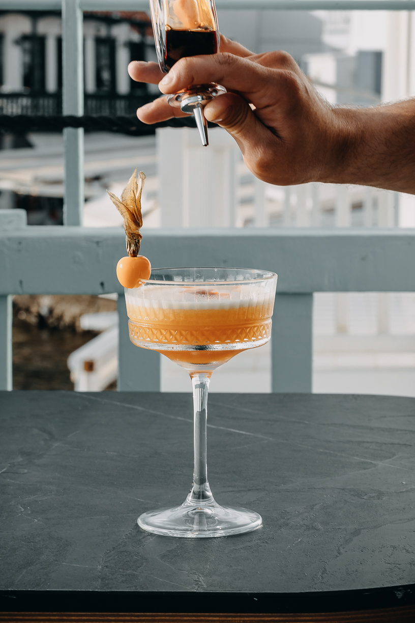 A coupe of amber cocktail garnished with a physalis, held against the sea.
